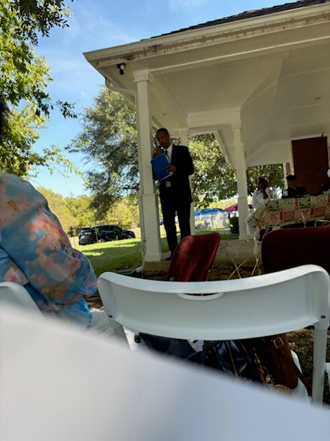 A speaker holding a blue folder addresses the gathering from the porch.