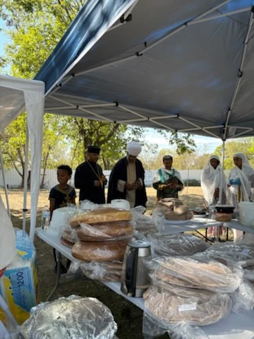 Clergy and community members gather beside a table of food beneath an outdoor canopy.