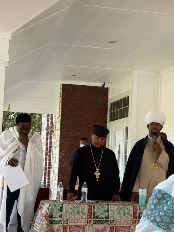 Three clergy members stand behind a covered table on the porch, with one speaking into a microphone.