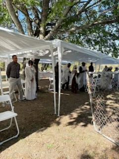 Community members gather under white canopies among trees on the monastery grounds.
