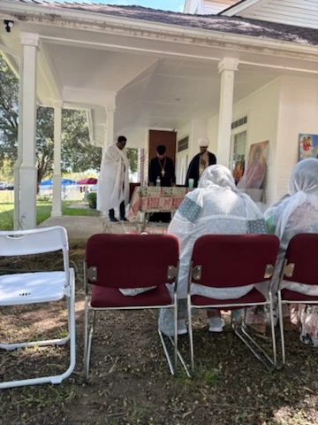 Members seated outdoors face clergy gathered at a table on the residence porch.