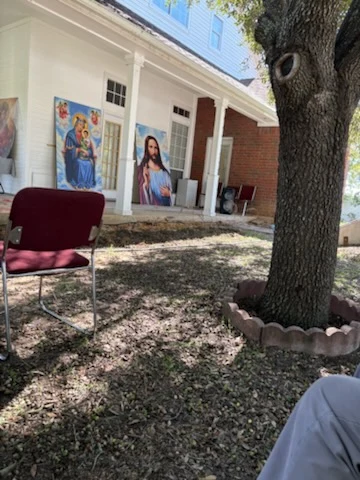 Sacred images stand along the residence porch beside chairs and a large shade tree.