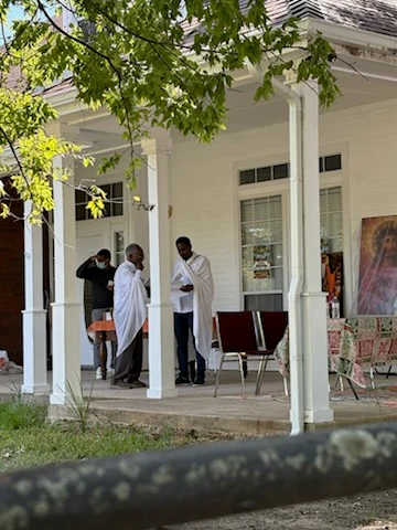 Two participants stand with papers on the shaded porch during the gathering.